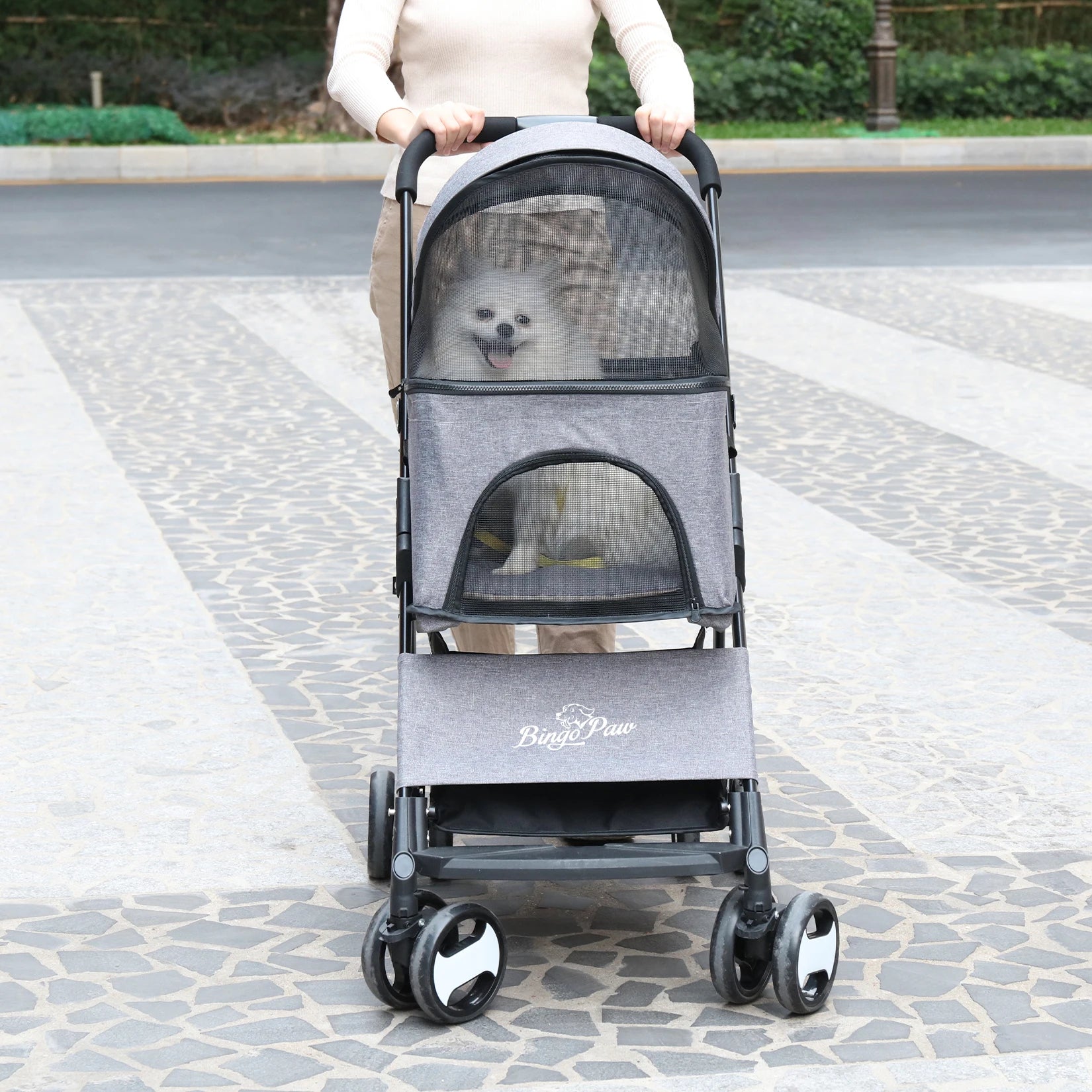 Person pushing a gray pet stroller with a small white dog inside on a paved walkway.