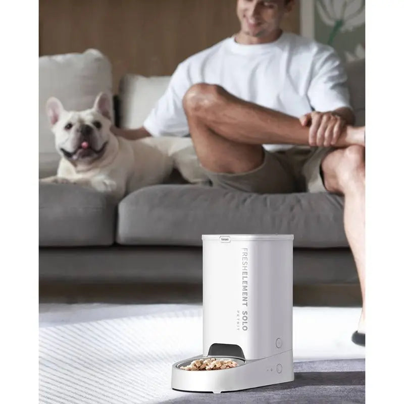 Person sitting on a couch with a dog, and a white pet feeder on the floor.