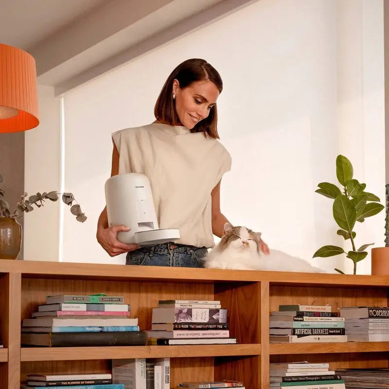 Woman holding a white appliance next to a cat on a bookshelf with books and plants.