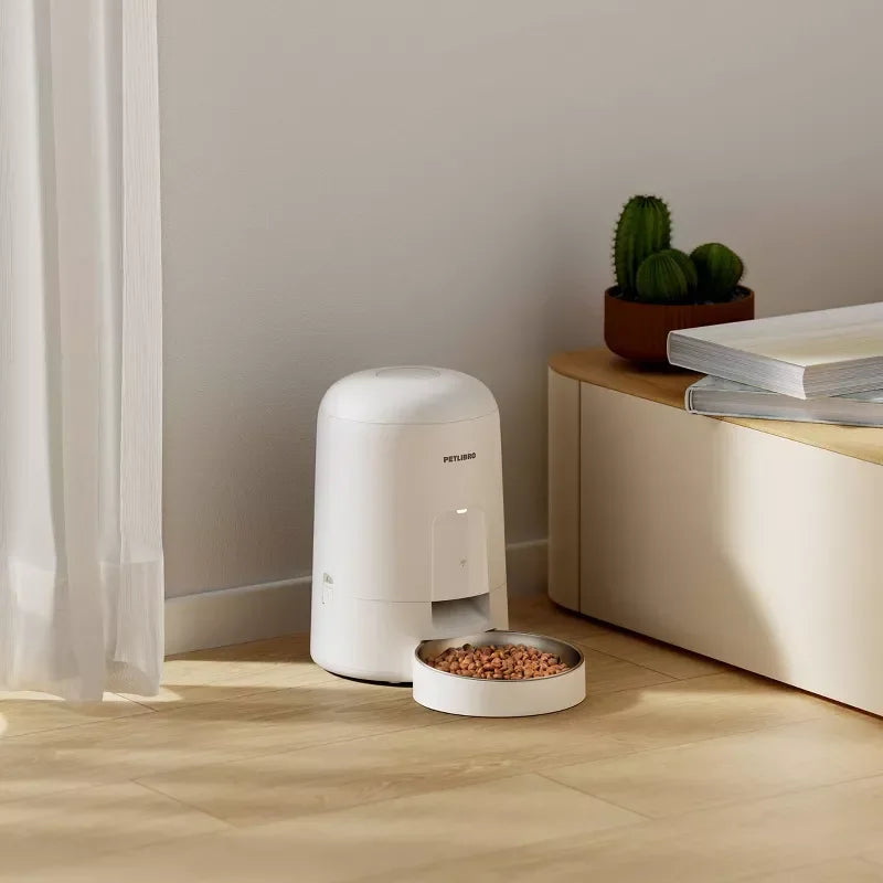 White pet feeder on a wooden floor with a bowl of food, next to a curtain and a small table with books and a plant.